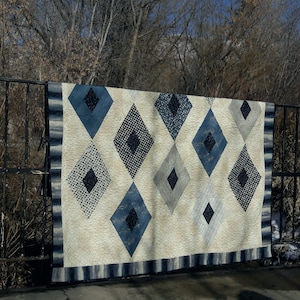 May include: A white quilt with a diamond pattern in shades of blue and black. The quilt is hanging on a fence.
