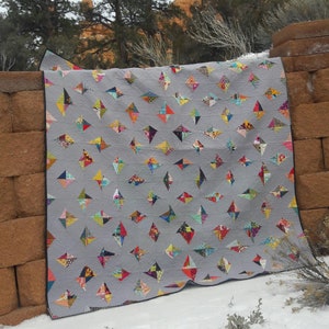 May include: A gray quilt with a diamond pattern made from colorful fabric scraps. The quilt is hanging on a brick wall with a snowy background.