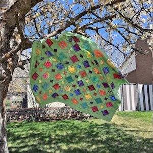 May include: A colourful quilt with a diamond pattern hangs from a tree branch. The quilt is made of various fabrics in shades of green, red, yellow, blue, and purple. The quilt is hanging in a backyard with green grass and a tree in the background.