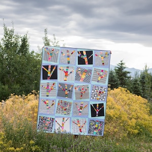 Op de afbeelding: Een kleurrijk quilt met een patchwork-ontwerp met meerdere bomen met takken gemaakt van verschillende gekleurde stoffen. De quilt wordt getoond tegen een achtergrond van een bewolkte lucht en groen gebladerte.