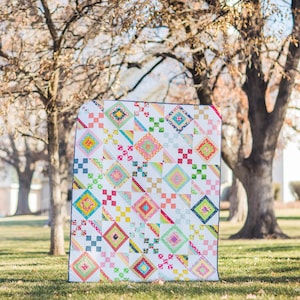 Op de afbeelding: Een kleurrijk patchwork-deken met een witte achtergrond en een verscheidenheid aan felle kleuren, waaronder roze, geel, blauw en groen. Het deken is gemaakt van vierkanten en ruiten en wordt tentoongesteld op een gazon.