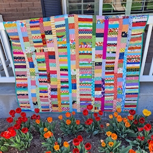 May include: A colorful patchwork quilt with a variety of fabrics and patterns. The quilt is made up of horizontal strips of fabric, each with a different color and design. The quilt is hanging on a wall and is surrounded by a bed of red tulips.