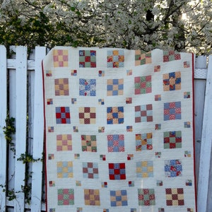 May include: A handmade quilt with a cream-colored background and a patchwork design of squares in various colors and patterns, including red, blue, yellow, and green. The quilt is hanging on a white picket fence, with a red border.