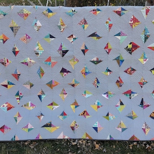 以下が含まれることがあります： グレーの背景にカラフルなダイヤモンドの形のパターンが施されたキルト。キルトは、さまざまなパターンと色の異なる生地で作られています。