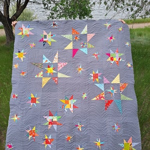 Könnte beinhalten: Eine handgefertigte Steppdecke mit grauem Hintergrund und Wellenmuster. Sie zeigt zahlreiche bunte Sternformen in verschiedenen Größen, mit einem Patchwork-Design aus leuchtenden Stoffen. Die Steppdecke wird von unsichtbaren Händen gehalten.