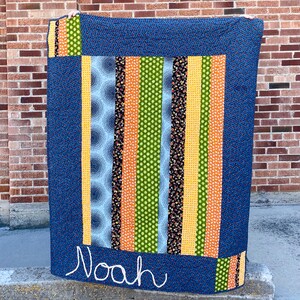May include: A blue and yellow striped quilt with a name, "Noah", embroidered on the bottom. The quilt is made of various fabrics with different patterns, including circles, stripes, and flowers.