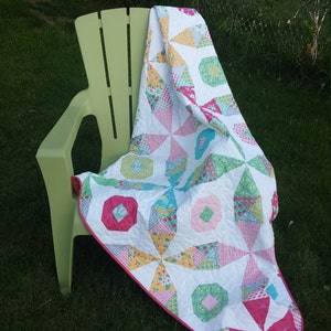 May include: A white quilt with a pink, green, and yellow floral pattern is draped over a green plastic chair. The chair is sitting on a grassy lawn.