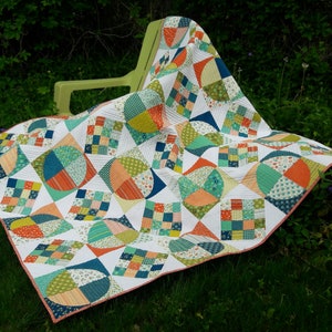 May include: A colorful patchwork quilt with a geometric design featuring circles, squares, and triangles. The quilt is made with a variety of fabrics in shades of blue, green, orange, and white. The quilt is draped over a green chair and is sitting on a bed of green grass.