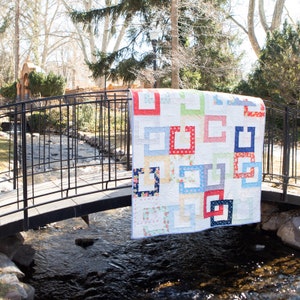 Peut inclure: Une courtepointe faite à la main avec un fond blanc et des motifs carrés dans des carrés colorés en rouge, bleu, vert et jaune. La courtepointe est drapée sur un pont en métal noir, avec un ruisseau et des arbres en arrière-plan.