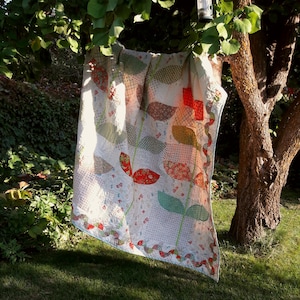 May include: A white quilt with a floral pattern in shades of red, green, and blue. The quilt is hanging on a tree branch and is being illuminated by the sun.