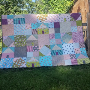 May include: A colourful patchwork quilt with a variety of patterns and colours, including pink, blue, green, and brown. The quilt is made up of squares and triangles, and it features a design of houses.