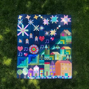 May include: A handmade quilt featuring a whimsical town scene with houses, trees, stars, and musical notes. The quilt is primarily navy blue with colorful patchwork details. The quilt is displayed on a green grassy surface.