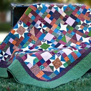 May include: A colourful patchwork quilt with a variety of geometric shapes and star patterns. The quilt, with a green border, is draped over a dark trunk. The quilt's design incorporates blue, green, orange, and purple hues.