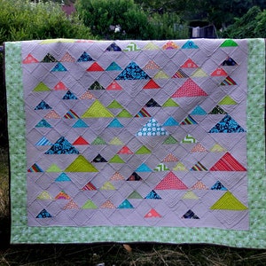 May include: A patchwork quilt featuring a repeating pattern of colourful triangles on a grey background. The triangles are in various patterns and shades of blue, green, red, and orange. The quilt has a green border and is displayed on a chain-link fence.