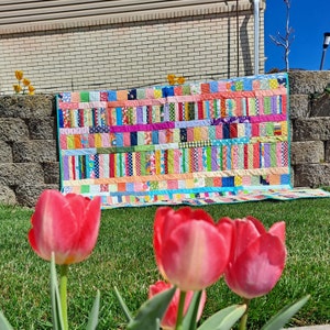 May include: A colorful patchwork quilt with a variety of fabrics and patterns. The quilt is made up of strips of fabric in different colors and designs, including pink, blue, green, yellow, and red. The quilt is laid out on the ground in front of a brick wall.