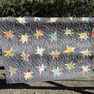 Könnte beinhalten: Eine handgefertigte Steppdecke mit grauem Hintergrund und einem Muster aus bunten Sternen. Die Sterne bestehen aus verschiedenen gemusterten Stoffen in Rot-, Gelb-, Blau- und Grüntönen. Die Steppdecke wird im Freien präsentiert.