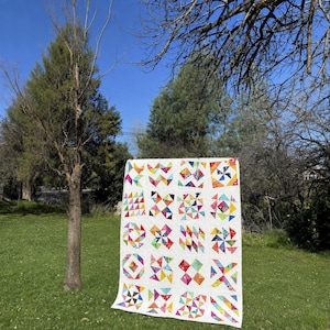 Puede incluir: Una colcha rectangular con fondo blanco y una cuadrícula de bloques geométricos coloridos y estampados. Los bloques muestran varias formas, incluyendo triángulos, molinillos y cuadrados en tonos vibrantes de rosa, amarillo, azul y verde.