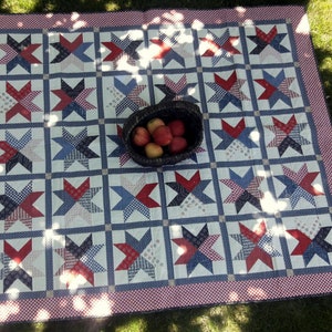May include: A handmade quilt with a star pattern in red, white, and blue. The quilt is laid out on the grass, with a basket of apples in the center. The border is a red and white gingham pattern.