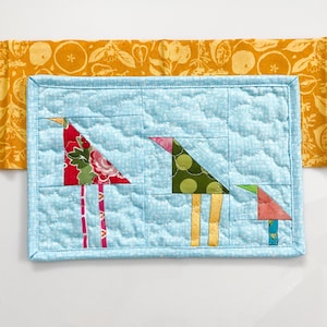 May include: A blue quilted hot pad with three colorful birds made from fabric scraps. The birds have long legs and are standing on the pad.