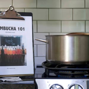 Pode incluir: Uma panela de aço inoxidável com uma colher de pau em cima está em cima de um fogão preto. Uma prancheta com o texto "Kombucha 101" está em uma bancada ao lado do fogão. Também há uma xícara medidora e um saquinho de chá na bancada.