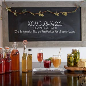 May include: A chalkboard sign with the text "Kombucha 2.0 Beyond the Brew 2nd fermentation Tips and Fun Recipes For all Booch Lovers" with a variety of kombucha bottles, jars of pickled vegetables, and glasses of kombucha on a wooden table.