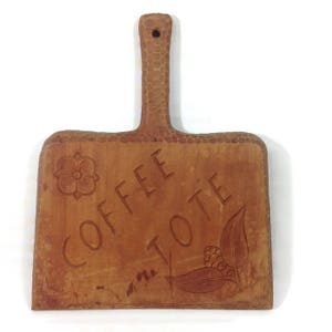 May include: A wooden chopping board with a carved handle and the words "Coffee Tote" carved into the surface. The board is brown and has a rustic look.