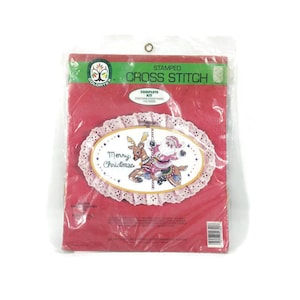 May include: A red and white package containing a cross stitch kit with a pink and white oval frame. The frame has a cross stitch design of a reindeer pulling a carousel with Santa Claus on it. The text "Merry Christmas" is stitched in the frame. The package says "Stamped Cross Stitch Complete Kit Contains Everything You Need".