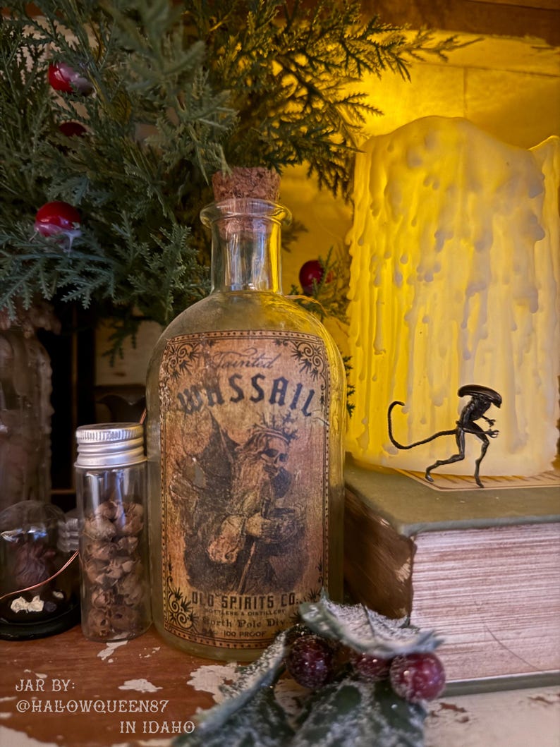 Pu&ograve; includere: Una bottiglia in vetro trasparente con tappo in sughero e un'etichetta in stile vintage con la scritta Tainted WASSAIL, OLD SPIRITS CO., North Pole Div., 100 PROOF, raffigurante una figura scheletrica con corona e un bastone.