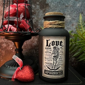 May include: A matte black bottle with a cork stopper and twine wrapped around the neck. The label features a skeleton illustration and text reading Love Potion No. 13, Stimulant of Desire, Extra Passion, and Aphrodite Brand in black and white.