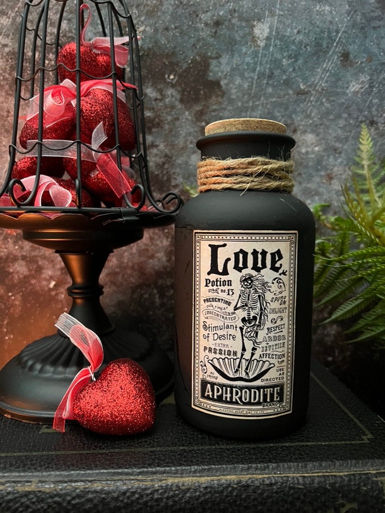 May include: A matte black bottle with a cork stopper and twine wrapped around the neck. The label features a skeleton illustration and text reading Love Potion No. 13, Stimulant of Desire, Extra Passion, and Aphrodite Brand in black and white.