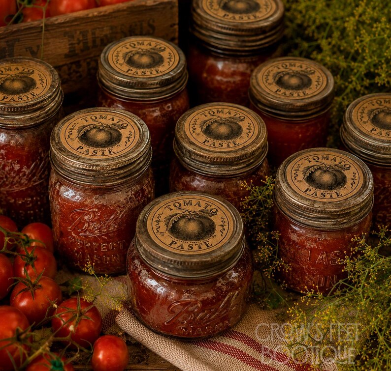 Op de afbeelding: Verschillende glazen potten gevuld met rode tomatenpuree, elk voorzien van een metalen deksel en een rond beige etiket met een tomatenillustratie en de tekst Tomato Paste. Verse rode tomaten en groene dille zijn in de buurt gerangschikt.