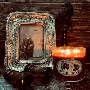 May include: A three-wick candle in a clear glass jar with a label featuring two hanging bats and the text FOREVER TOGETHER, positioned next to a vintage-style silver ornate picture frame containing a black and white portrait and brass binoculars.