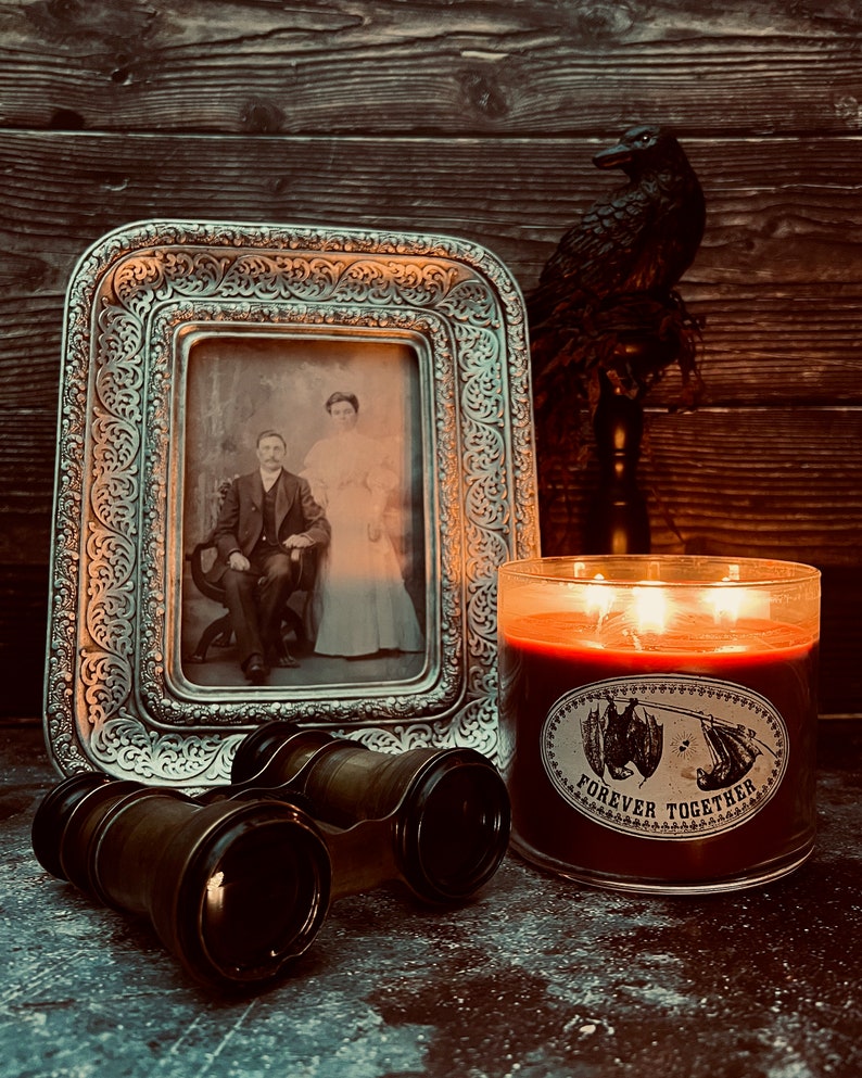May include: A three-wick candle in a clear glass jar with a label featuring two hanging bats and the text FOREVER TOGETHER, positioned next to a vintage-style silver ornate picture frame containing a black and white portrait and brass binoculars.