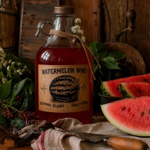 Op de afbeelding: Een heldere glazen kan gevuld met rode watermeloenwijn, voorzien van een beige etiket met een zwarte watermeloenillustratie en de tekst Watermelon Wine, Rush Springs, Oklahoma, Jubilee Wineries. Een touwtje en kleine witte bloemetjes sieren de hals.