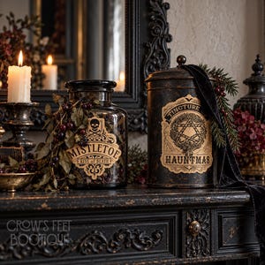 Pu&ograve; includere: Un barattolo di vetro trasparente etichettato MISTLETOE KISS OF DEATH con un'icona a teschio e un contenitore in metallo nero etichettato TINCTURE OF HAUNTMAS con un disegno a pentagramma. Entrambi sono decorati con fogliame scuro e bacche.