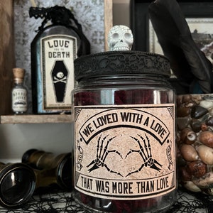 May include: A clear glass jar with a black lid topped by a small white skull. The jar features a label with the text "WE LOVED WITH A LOVE THAT WAS MORE THAN LOVE" and an illustration of two skeleton hands forming a heart shape in the center.