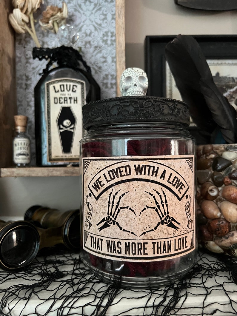 May include: A clear glass jar with a black lid topped by a small white skull. The jar features a label with the text "WE LOVED WITH A LOVE THAT WAS MORE THAN LOVE" and an illustration of two skeleton hands forming a heart shape in the center.