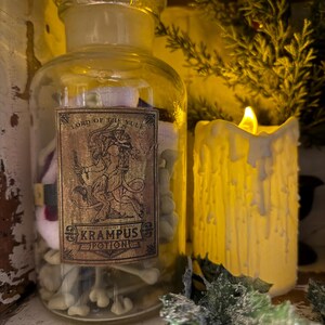 Pu&ograve; includere: Un barattolo da farmacia in vetro trasparente con tappo in vetro, contenente piccoli oggetti bianchi a forma di osso. Un'etichetta rettangolare sul davanti presenta un'illustrazione di Krampus e il testo: Lord of the Yule, Krampus Potion.
