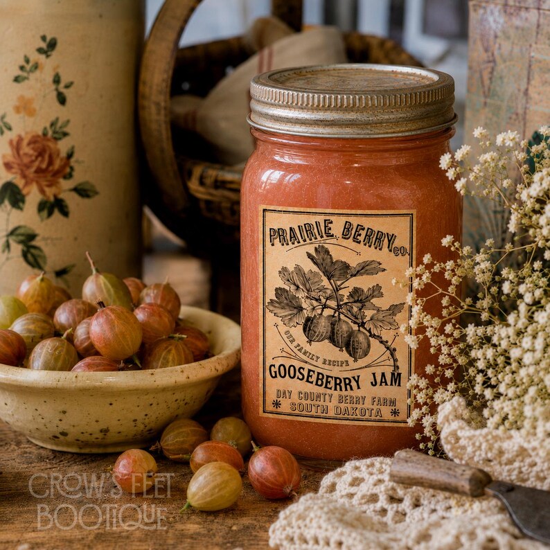 Op de afbeelding: Een glazen pot gevuld met oranje kruisbessenjam met een etiket waarop staat Prairie Berry Co. Gooseberry Jam, Our Family Recipe, Day County Berry Farm, South Dakota. Een kom met verse kruisbessen staat naast de pot.