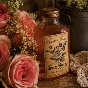 Op de afbeelding: Een doorzichtige glazen fles gevuld met roze rozenwater, voorzien van een bruin rechthoekig etiket met zwarte tekst Summer Garden, A Cosmetic Marvel, Rose Water, Adelin Edith Lewis Co. en een gedetailleerde zwart-witte roosillustratie.