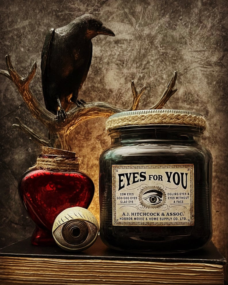 May include: A dark glass jar with a label reading EYES FOR YOU, A.J. HITCHCOCK & ASSOC. HORROR MOVIE & HOME SUPPLY CO. LTD., featuring a central eye illustration. Beside it sits a red heart-shaped bottle, a spherical eye prop, and a black raven figure.