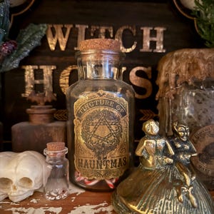 Pu&ograve; includere: Un barattolo da farmacia in vetro trasparente con tappo in sughero, dotato di un'etichetta dorata con la scritta Tincture of Hauntmas, riempito di piccoli bastoncini di zucchero. Il barattolo &egrave; circondato da una statuetta in ottone e un teschio.