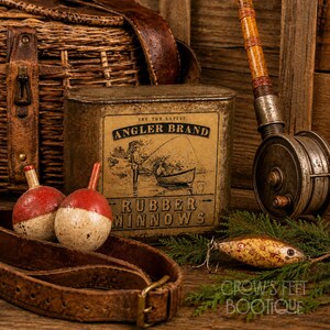 Op de afbeelding: Een vintage metalen blik met het label Angler Brand Rubber Minnows met een afbeelding van een persoon die vanuit een boot vist. Het blik is omringd door twee ronde rood-witte visdobbers, een hengel met een molen en een leren riem.
