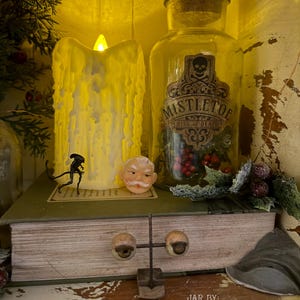 Pu&ograve; includere: Una candela a pilastro LED gialla con effetto cera gocciolante, un barattolo di vetro trasparente etichettato MISTLETOE KISS OF DEATH con un'icona a teschio, una piccola statuetta aliena, un ornamento a faccia di Babbo Natale e un supporto in metallo.