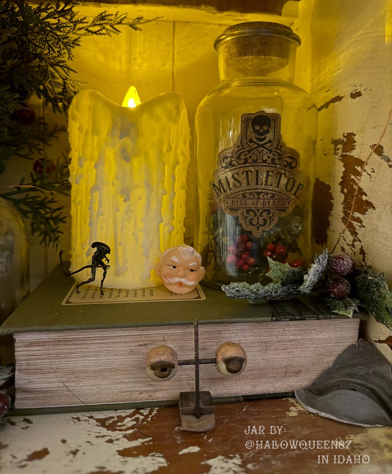 Pu&ograve; includere: Una candela a pilastro LED gialla con effetto cera gocciolante, un barattolo di vetro trasparente etichettato MISTLETOE KISS OF DEATH con un'icona a teschio, una piccola statuetta aliena, un ornamento a faccia di Babbo Natale e un supporto in metallo.