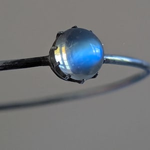 Pode incluir: Uma pulseira de metal prateado com uma única pedra da lua redonda de corte cabochão em uma cravação de garras. A pedra da lua apresenta uma aparência translúcida e leitosa com uma distinta adularescência azul em sua superfície.