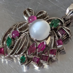 Puede incluir: Un collar con colgante de plata con una piedra central blanca y redonda, rodeada de piedras rojas y verdes. El colgante tiene un diseño floral con un borde de cuerda trenzada y está unido a una cadena de plata.