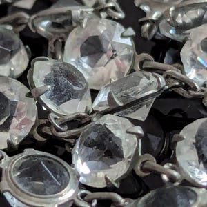 Op de afbeelding: Close-up van een zilverkleurige ketting met grote, facetgeslepen, heldere, kristalachtige stenen. De stenen zijn verbonden met zilverkleurig metaal. De donkere achtergrond creëert contrast.