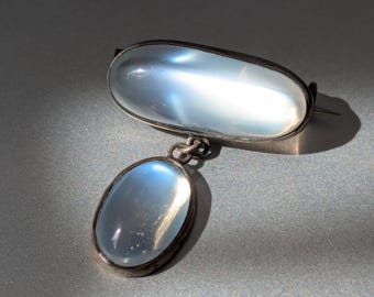 Könnte beinhalten: Eine silberfarbene Brosche mit einem großen, horizontalen, ovalen Cabochon-Stein mit einem durchscheinenden, milchig-blauen Schimmer. Ein kleinerer, vertikaler, ovaler Cabochon-Stein mit gleichem Aussehen hängt an einem Metallring am Hauptstück.