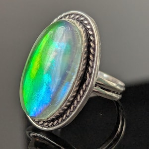 May include: A silver ring with an oval, iridescent gemstone displaying green, blue, and yellow colours. The gemstone is set in a silver bezel with a rope-style border. The ring has a double-banded shank.